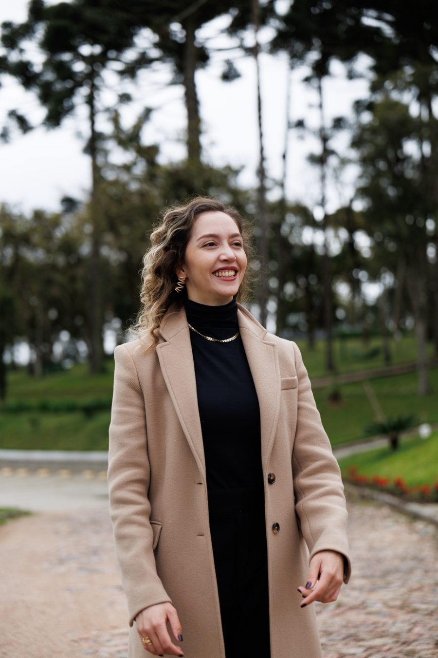 Deyse Schultz sorrindo ao ar livre, de casaco bege, com araucárias ao fundo